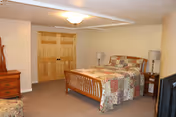 A bedroom with a wooden bed frame and a colorful patchwork quilt. There are two matching bedside tables with lamps on each side of the bed. A wooden dresser is visible on the left side of the room. The walls are painted light beige, and there is a double wooden door in the background.