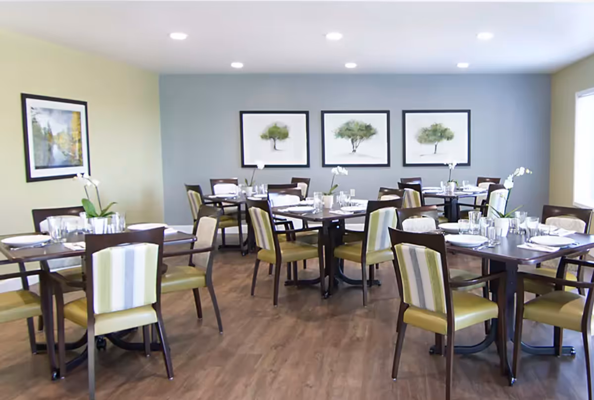 Dining room with several tables set for meals, green-upholstered chairs, and framed tree artwork on the wall.