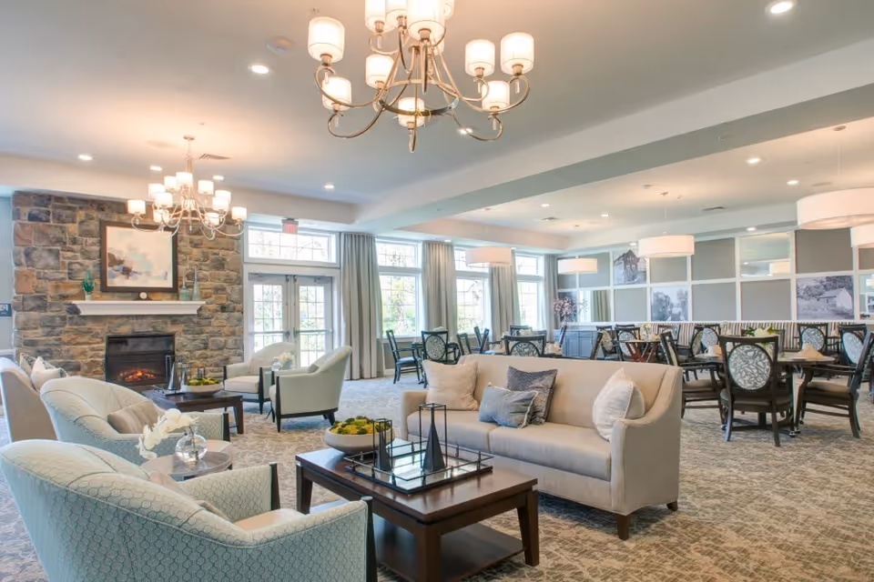 A spacious and well-lit common area with comfortable seating including armchairs and a sofa around a wooden coffee table. The room features a stone fireplace with a painting above it, large windows with curtains, and multiple chandeliers and pendant lights. In the background, there are several dining tables and chairs arranged neatly.