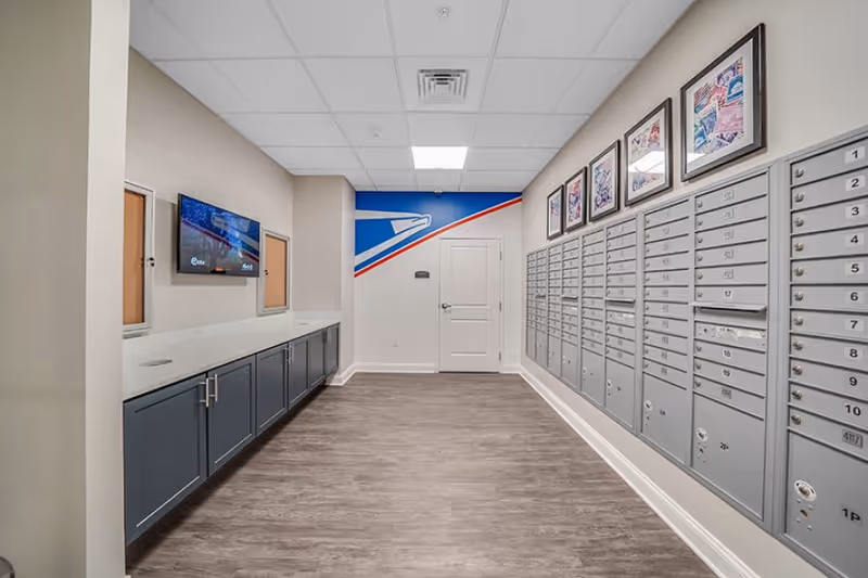 Interior view of a mailroom with multiple gray mailboxes mounted on the right wall, a white door at the end of the room, and a blue, red, and white mural featuring an eagle on the back wall. On the left side, there are blue cabinets with a white countertop, bulletin boards, and a wall-mounted TV screen. The floor is wood-patterned, and the ceiling has recessed lighting.