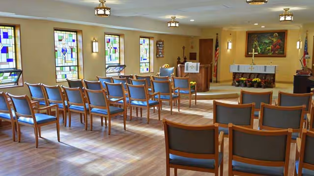 Interior view of a chapel with rows of wooden chairs with blue cushions facing an altar. The room has stained glass windows on the left wall, wall-mounted lights, and ceiling light fixtures. The altar area features a cross, floral arrangements, and a large framed painting on the back wall.