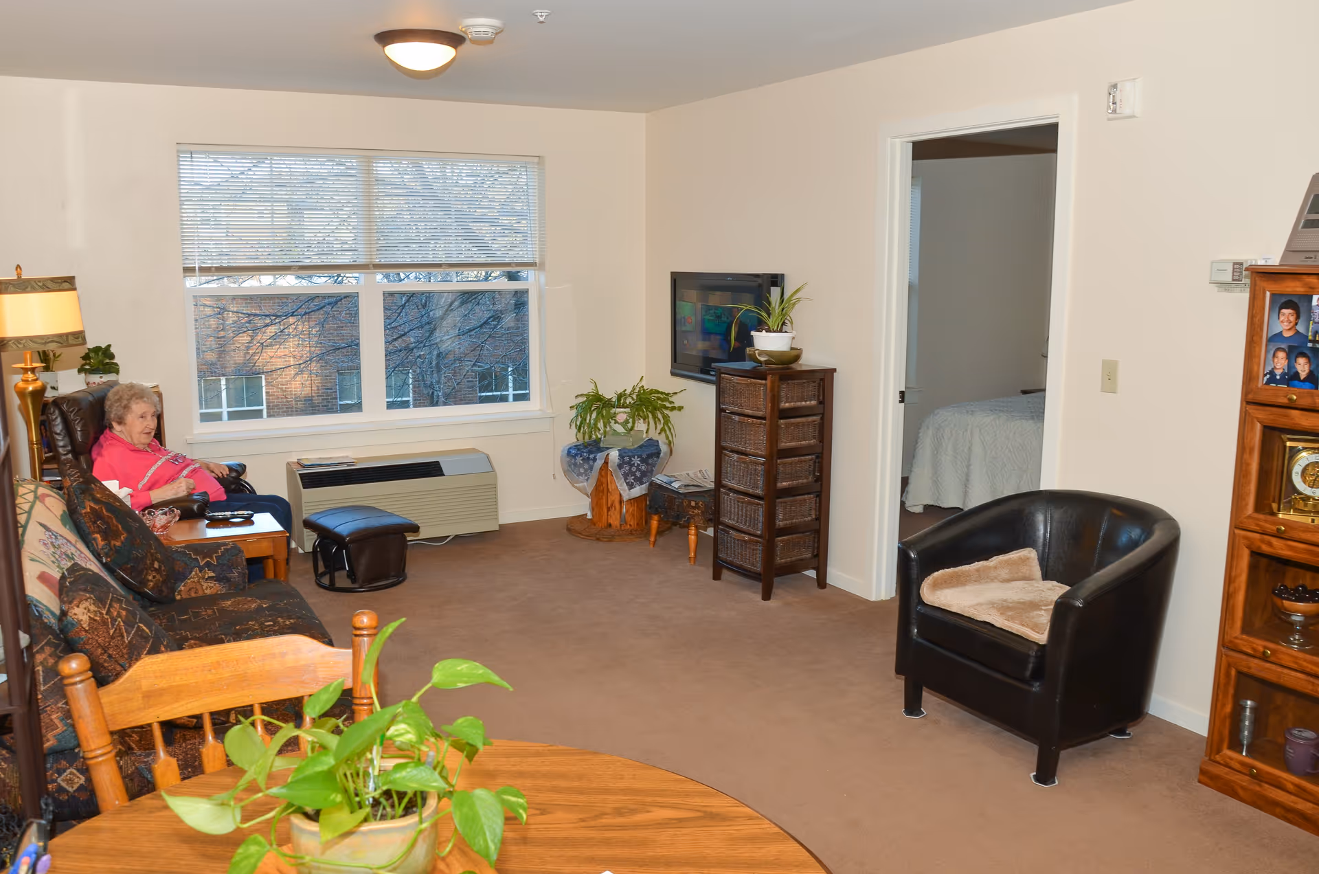 A cozy living room in a senior living facility with beige walls and carpet. There is a woman sitting on a patterned sofa near a window with blinds. The room contains a wooden rocking chair, a black leather armchair with a cushion, a small wooden table with a plant, a wicker drawer unit with a plant on top, and a wooden shelving unit with family photos and decorative items. A doorway leads to a bedroom with a bed covered in a white quilt.