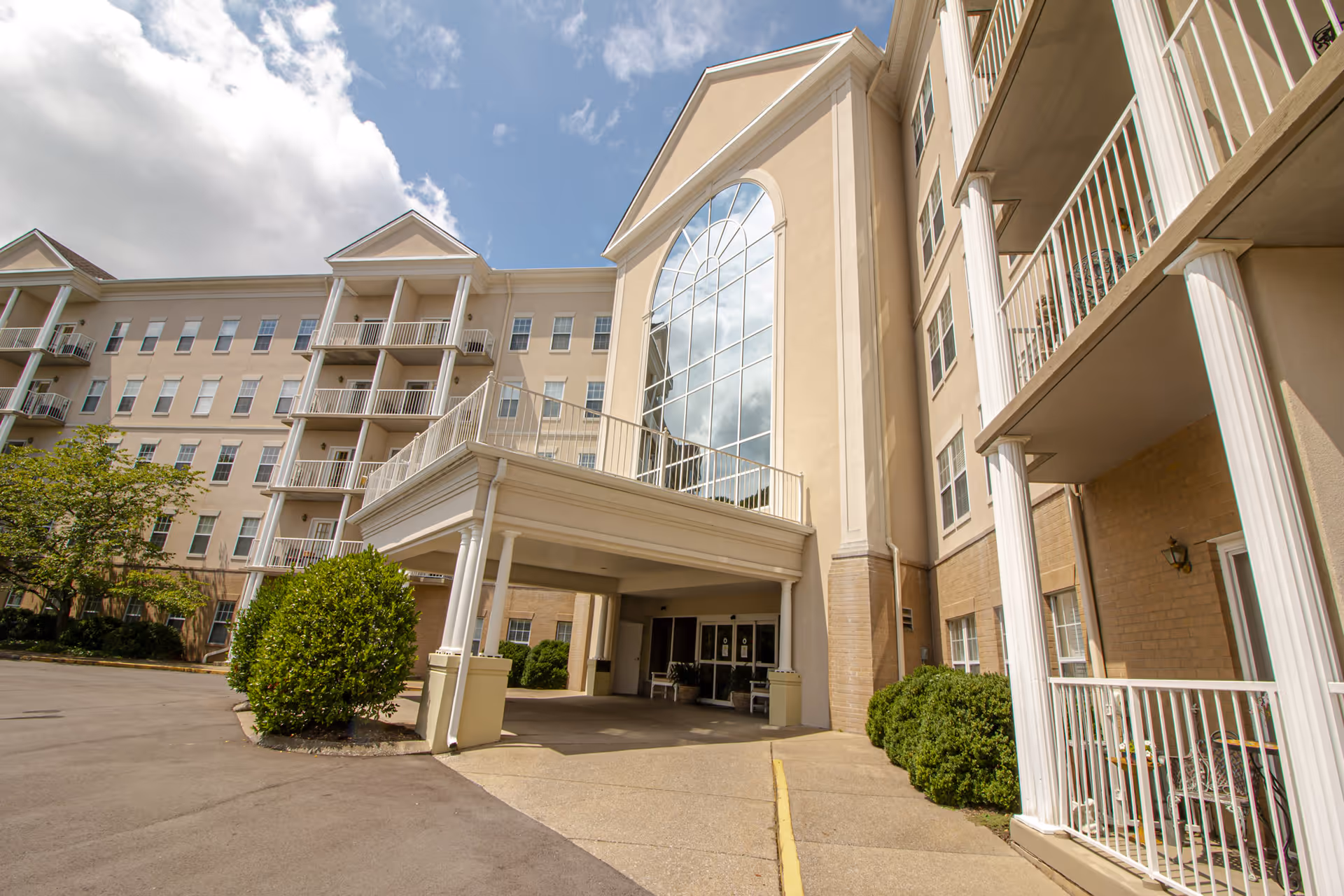 Exterior view of a multi-story senior living facility building with balconies, large windows, and a covered entrance under a partly cloudy sky.