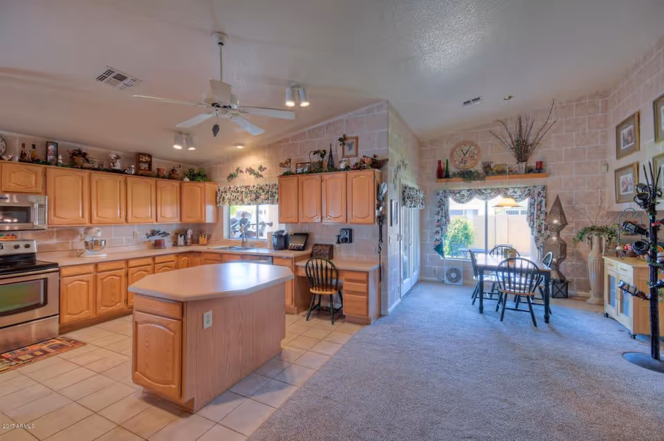 A spacious kitchen and dining area with wooden cabinets, a central island, and a dining table with chairs near a large window. The kitchen has a ceiling fan, stainless steel appliances, and decorative items on top of the cabinets. The dining area features carpeted flooring, wall art, and a large window with floral curtains letting in natural light.