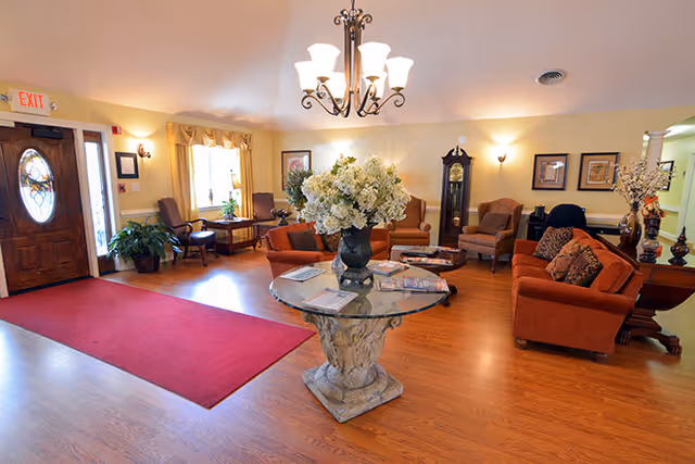Spacious senior living lounge with sofas and chairs around a central glass table topped with a large floral arrangement, a chandelier overhead, and the entrance door.