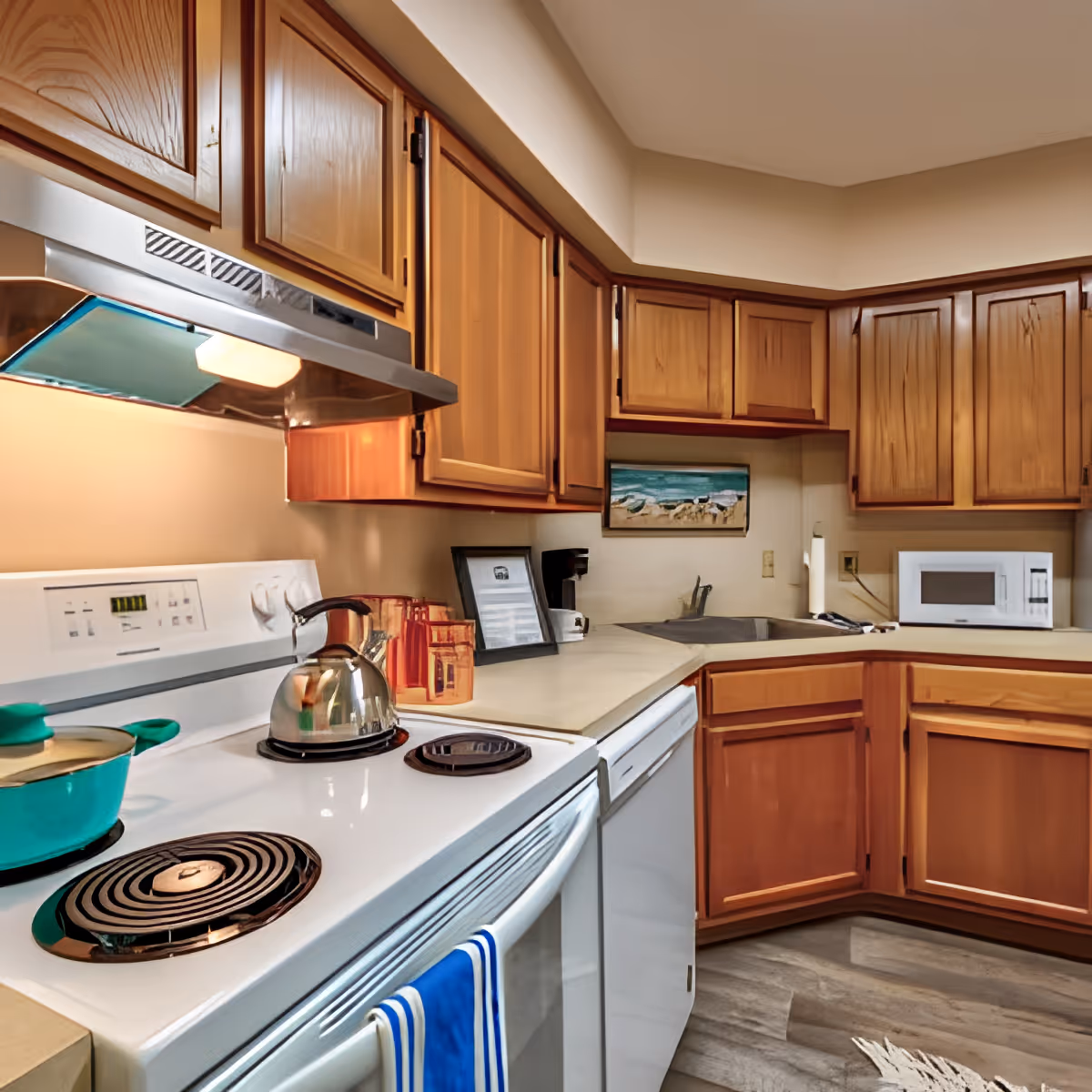 A kitchen with wooden cabinets, a white electric stove with a kettle and a pot on the burners, a dishwasher, a microwave, and a coffee maker. The countertop is light-colored, and there is a small framed picture and a painting of a beach scene on the wall.