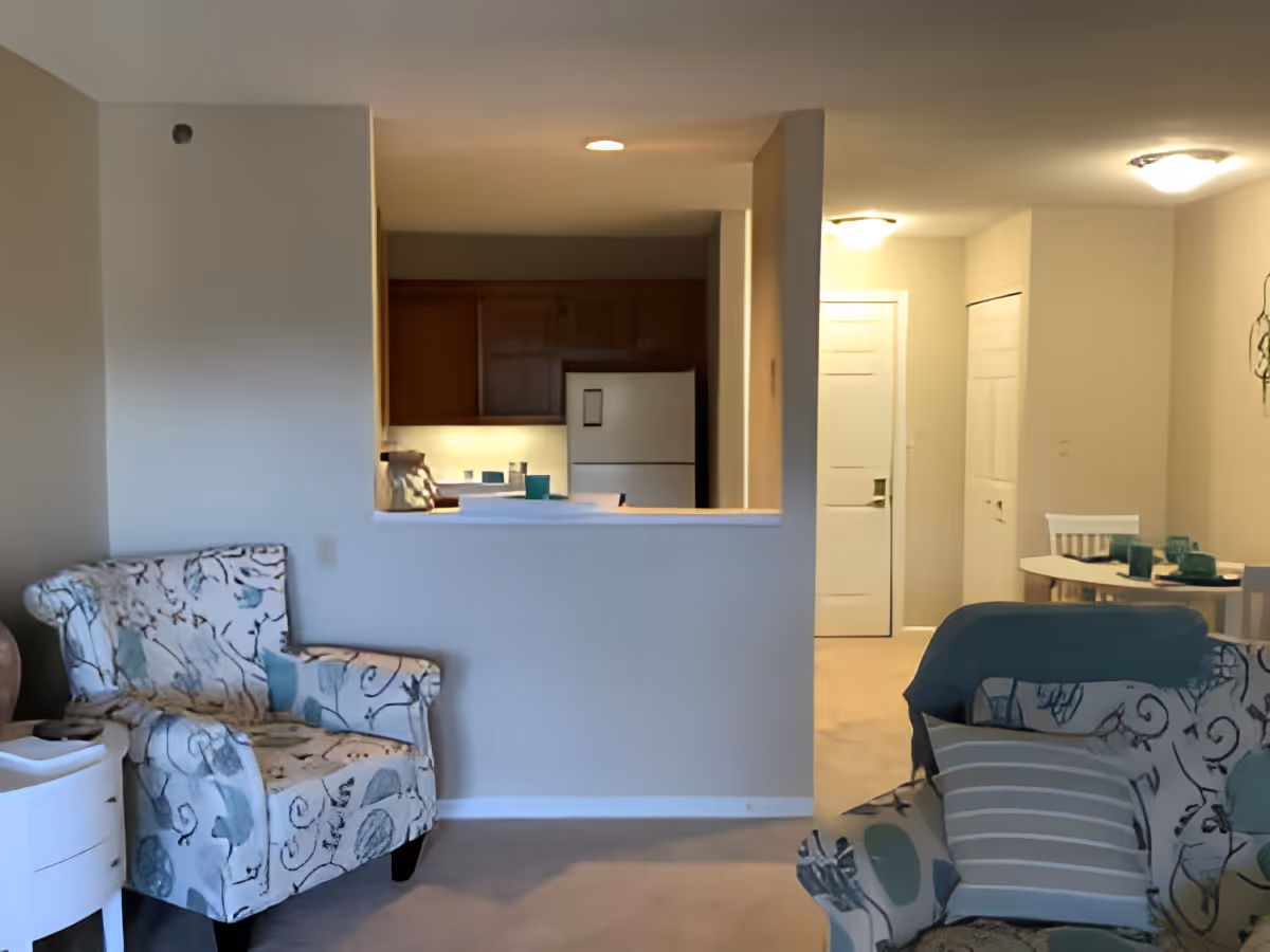 Interior view of a senior living facility showing a living room area with patterned armchairs and a small white side table. Beyond the living room is a kitchen with wooden cabinets and a white refrigerator, partially visible through a pass-through window. In the background, there is a dining area with a round table and chairs, set with teal-colored glassware. The walls are painted light beige, and the space is softly lit with ceiling lights.