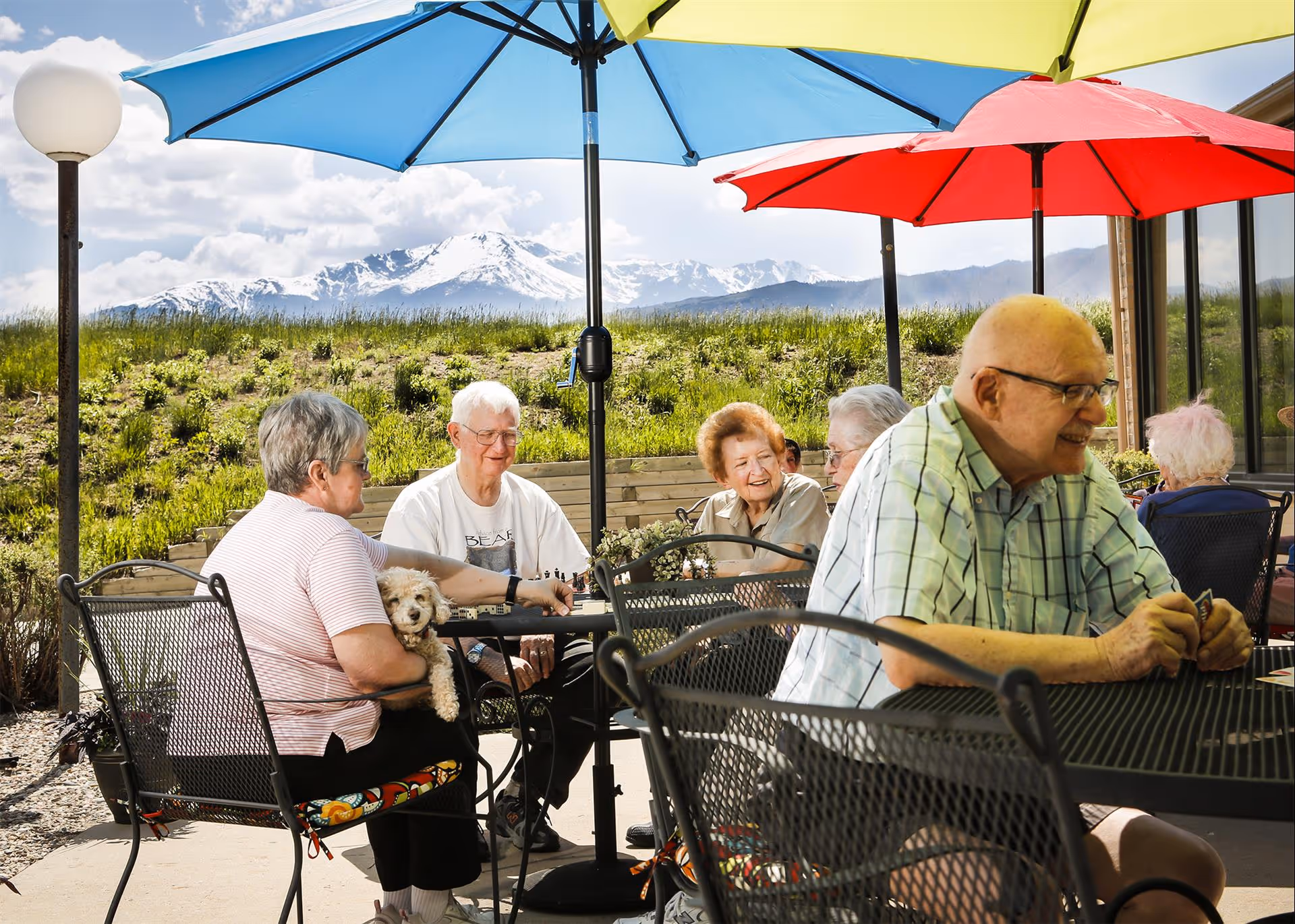 A group of elderly people sitting outside under colorful umbrellas at metal tables and chairs. One woman is holding a small dog. In the background, there is a grassy hill and snow-capped mountains under a partly cloudy sky.