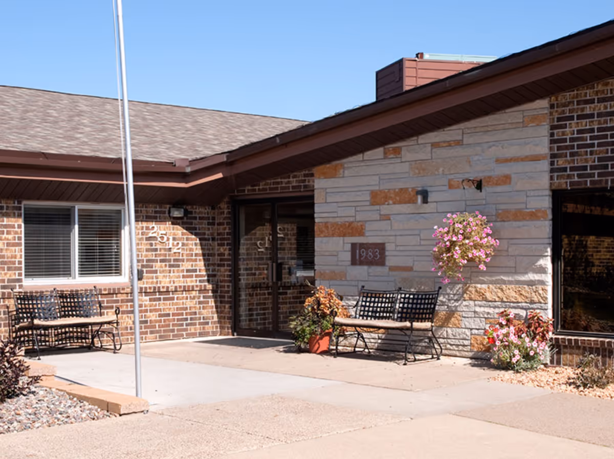 Front entrance of a brick senior living building with benches, potted flowers, hanging basket, and an address plaque reading 1983.
