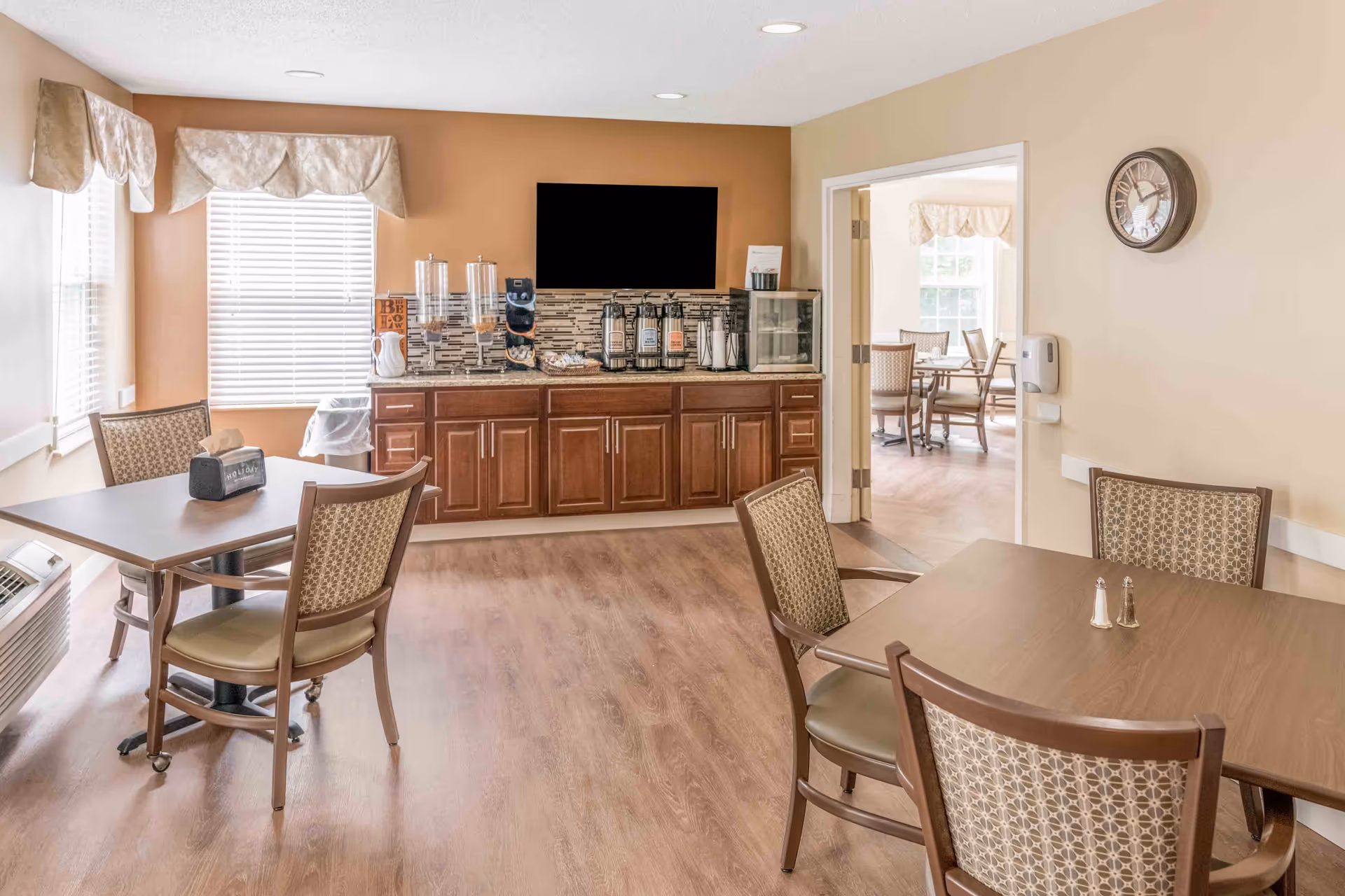 A bright dining area in a senior living facility with wooden tables and patterned chairs. There is a counter with coffee dispensers, cereal containers, and a small refrigerator against a wall with a mounted flat-screen TV. The room has large windows with valances and a wall clock, and an adjacent room with more dining tables is visible through an open doorway.