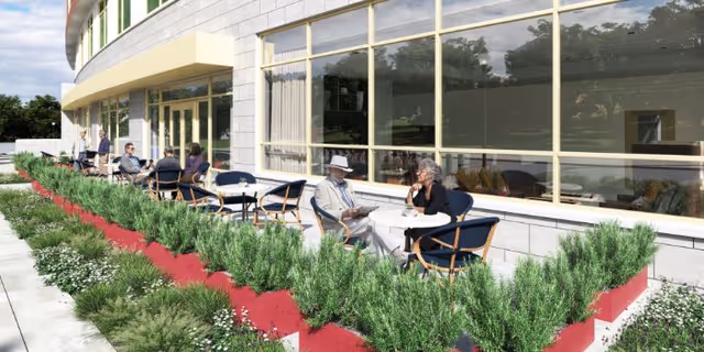Outdoor patio area at Benchmark at Alexandria with several elderly people sitting at round tables with chairs, enjoying the outdoor space. The patio is bordered by red planters filled with green shrubs and flowers. The building has large windows reflecting the surrounding greenery.