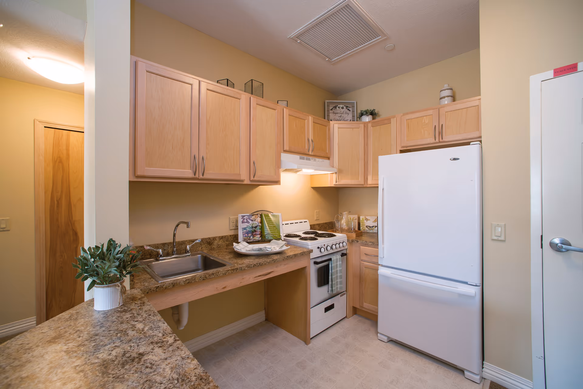 A small kitchen with light wood cabinets, a white refrigerator, a white stove with an oven, and a stainless steel sink. The countertops are brown with a speckled pattern. There are decorative items on top of the cabinets and a small plant on the counter. The walls are painted beige, and there is a door on the right side of the image.