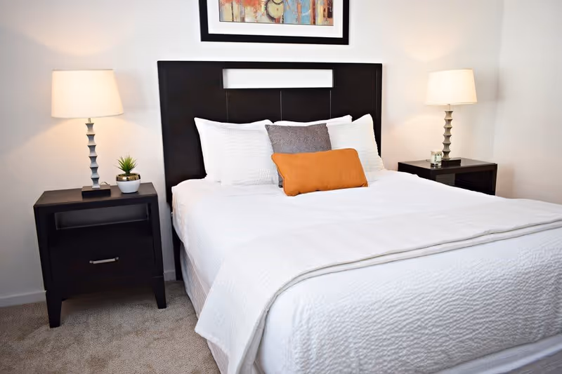 A neatly made bed with white bedding and three pillows, one gray and one orange, against a dark wooden headboard. On each side of the bed are black nightstands with modern table lamps and small decorative items. A colorful abstract painting hangs on the wall above the headboard.