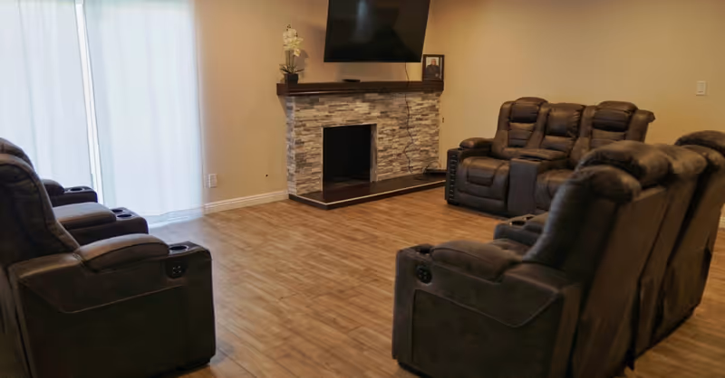A living room with several dark reclining chairs facing a wall-mounted TV above a stone fireplace and sliding glass doors.