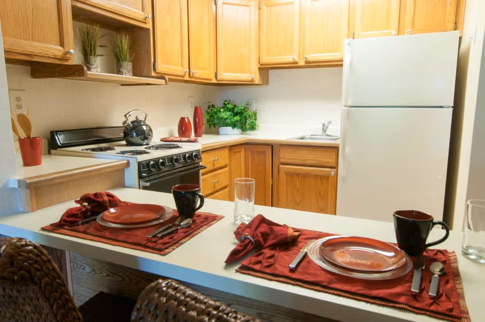 A small kitchen area with wooden cabinets, a white refrigerator, a stove with a kettle on it, and a countertop set for two with red placemats, plates, mugs, and utensils. There are decorative plants and red vases on the counter.