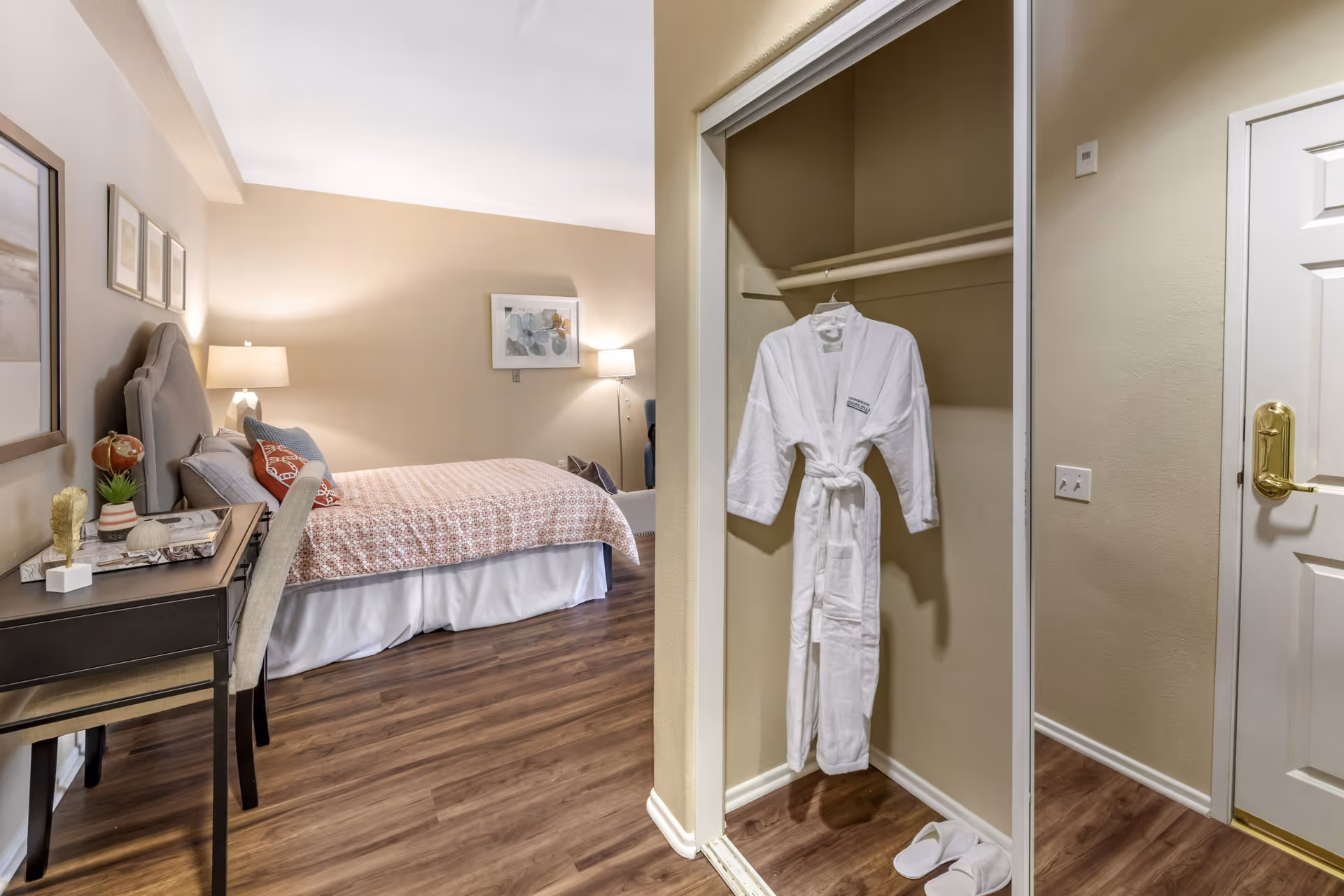 A cozy bedroom with a bed covered in a patterned bedspread, a bedside table with a lamp, and framed artwork on the wall. To the right, there is an open closet with a white bathrobe hanging inside and a pair of white slippers on the floor. The room has wooden flooring and beige walls.