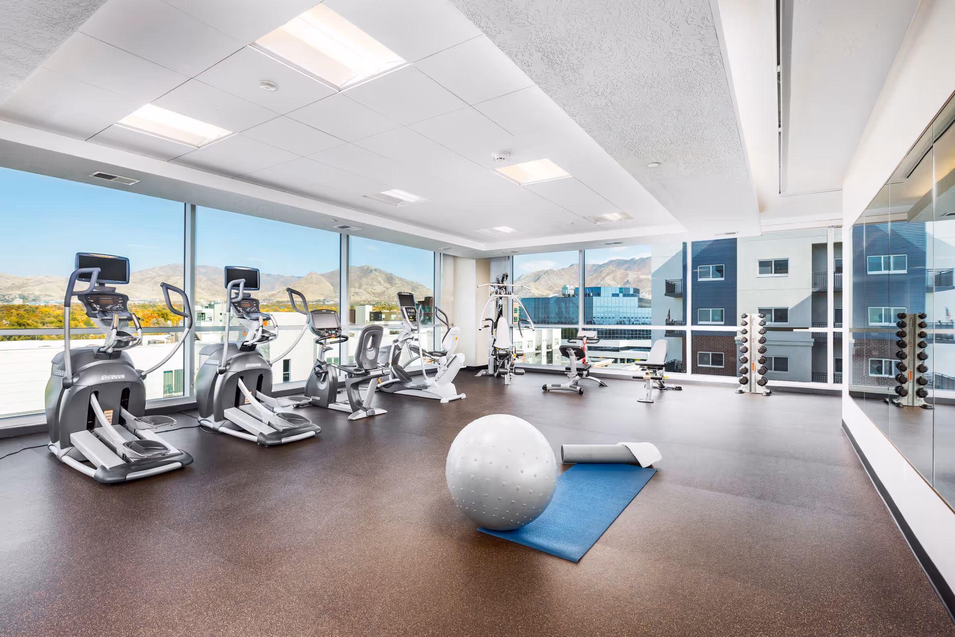 Bright and spacious fitness room with large windows offering a view of mountains and nearby buildings. The room contains several elliptical machines, stationary bikes, a multi-function weight machine, dumbbell racks, a large exercise ball, and a blue exercise mat with a foam roller. The floor is covered with brown rubberized material, and one wall is lined with mirrors.