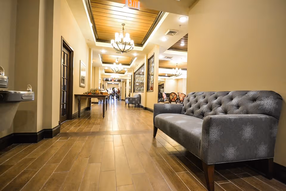 A well-lit hallway in a senior living facility with wooden floors and ceiling accents. There are chandeliers hanging from the ceiling, a gray tufted sofa on the right, a water fountain on the left, and a table with decorations further down the hall. People are visible in the distance near the end of the hallway.