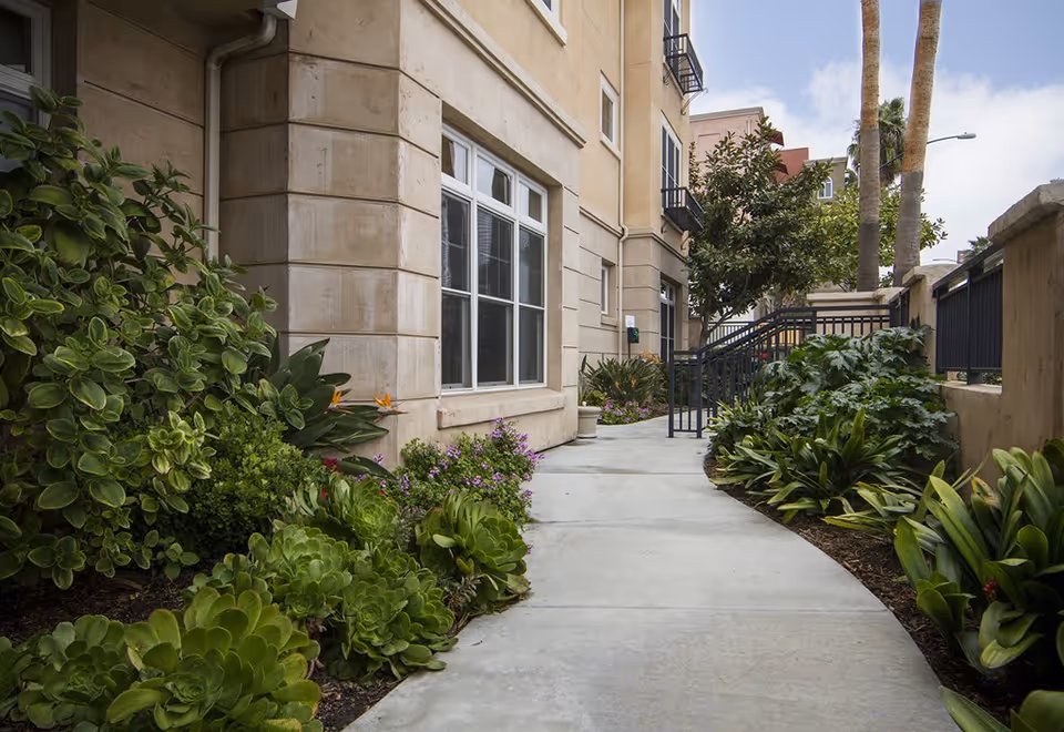 A concrete pathway lined with lush green plants and shrubs on both sides, leading alongside a beige building with multiple windows. There are palm trees and a black metal railing along the path, under a partly cloudy sky.