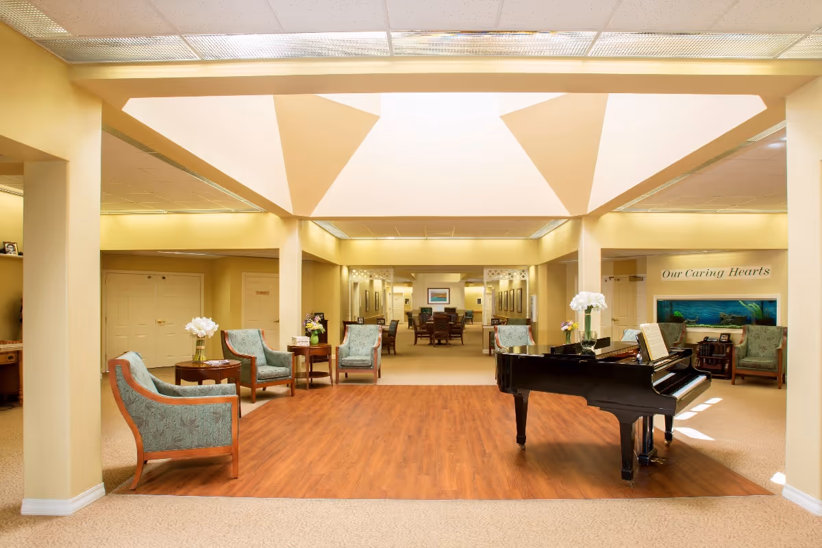 A spacious and well-lit common area in a senior living facility featuring a black grand piano on a wooden floor section, surrounded by several upholstered armchairs and small tables with flower arrangements. The walls are painted a soft yellow, and there is a large fish tank on the right side with the words 'Our Caring Hearts' displayed above it. The ceiling has a unique geometric design with recessed lighting.