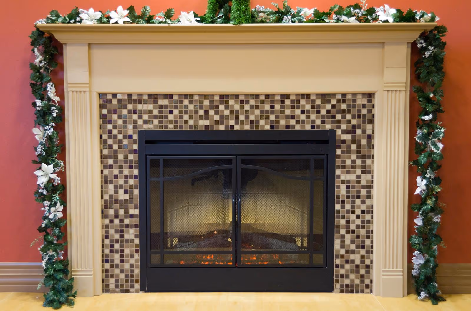 A decorative fireplace with a beige mantel and a mosaic tile surround in shades of brown and beige. The fireplace is adorned with a garland of green leaves and white flowers draped over the mantel and sides. The wall behind the fireplace is painted a warm reddish color, and the floor is light wood.