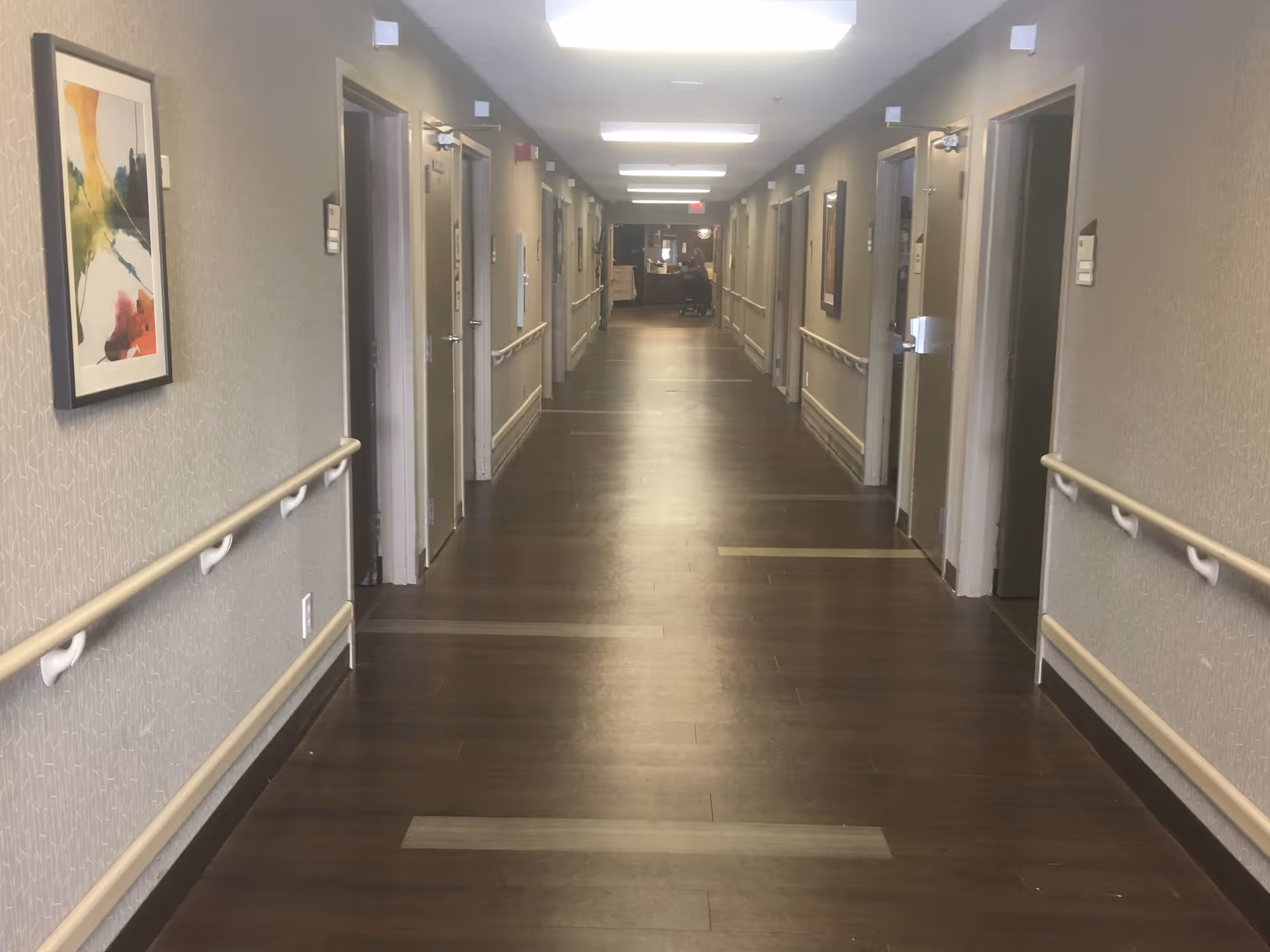 A long, well-lit hallway in a senior living facility with wooden floors, handrails on both sides, several closed doors, and framed artwork on the walls.
