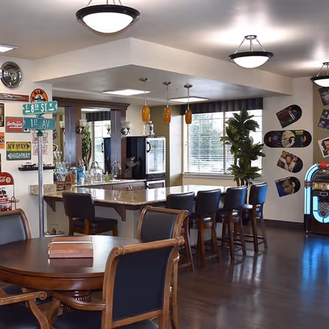Interior view of a senior living facility common area featuring a kitchen island with six bar stools, pendant lights hanging above, a round wooden table with chairs in the foreground, wall decorations including street signs and vinyl record covers, a jukebox, and large windows with blinds letting in natural light.
