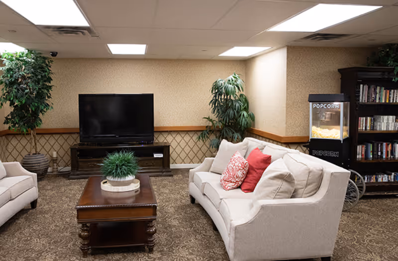 A cozy living room area with beige sofas adorned with red and patterned cushions, a wooden coffee table with a green plant centerpiece, a flat-screen TV on a wooden stand, two large potted plants, a popcorn machine, and a bookshelf filled with books and DVDs.
