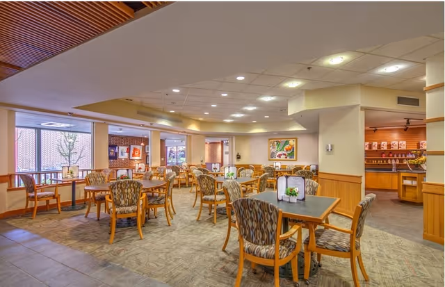 A spacious dining room with multiple wooden tables and patterned cushioned chairs arranged neatly. The room has large windows letting in natural light, framed artwork on the walls, and a serving area in the background. The ceiling has recessed lighting and the floor is carpeted with a patterned design.