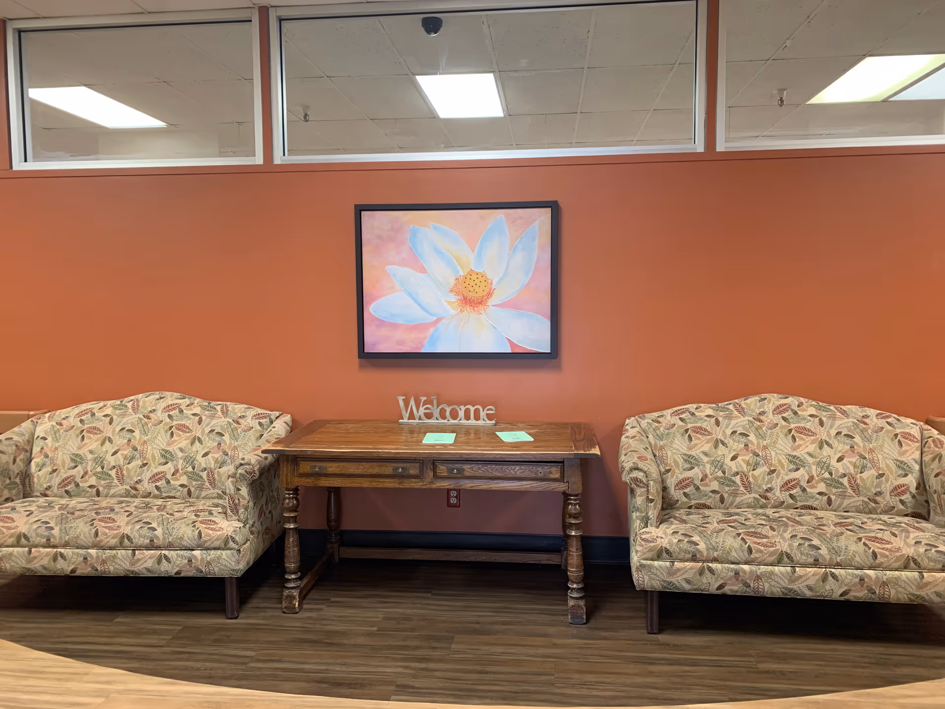 A seating area with two patterned upholstered loveseats facing each other, separated by a wooden table with a 'Welcome' sign and some papers on it. Above the table is a framed painting of a large white flower with a pink and orange background. The wall behind is painted a warm terracotta color, and there are windows near the ceiling.