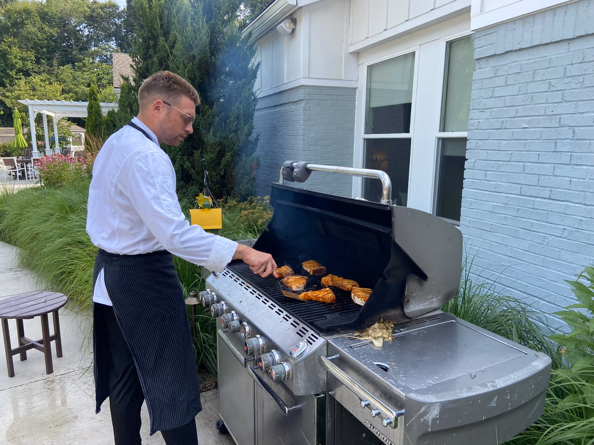 A man wearing a white chef's jacket and black apron is grilling several pieces of meat on a large outdoor gas grill next to a light blue brick building. There is greenery and outdoor furniture in the background.