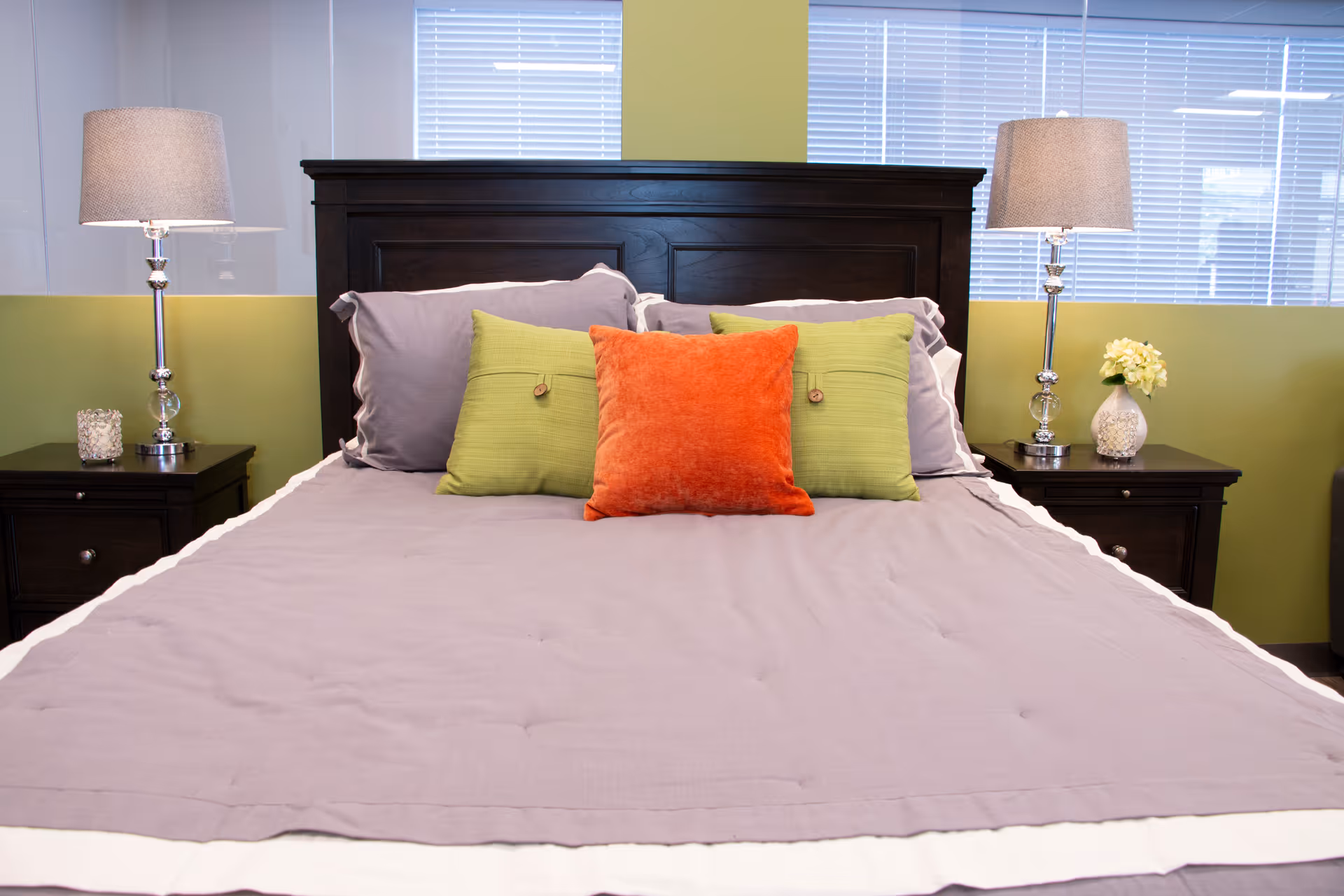 A neatly made bed with gray bedding, an orange and green accent pillows, a dark wood headboard and matching nightstands with lamps.