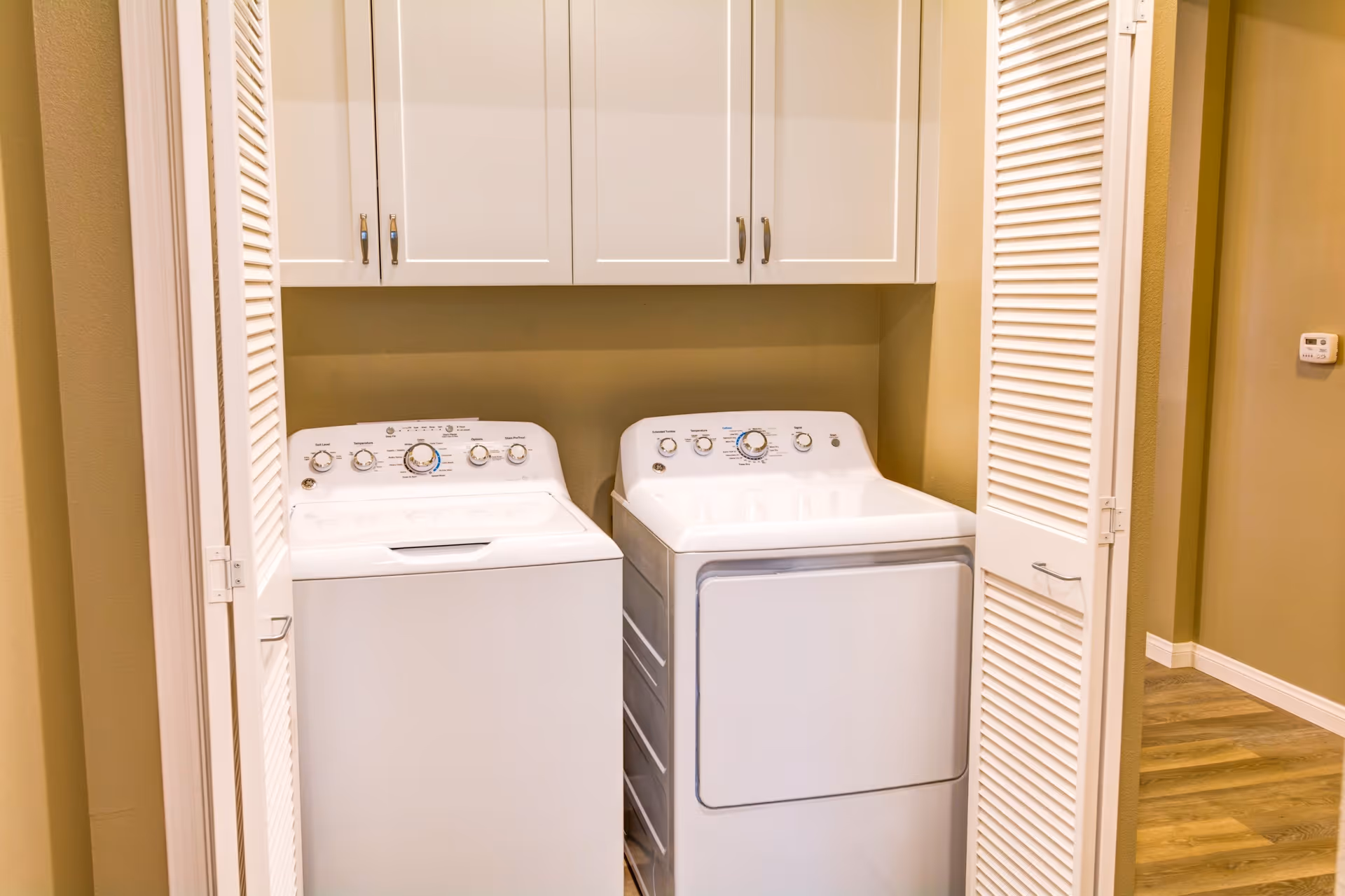 A laundry area with a white washing machine and dryer side by side, enclosed by white louvered doors. Above the machines are white cabinets for storage. The walls are painted beige and the floor has wood-like flooring.