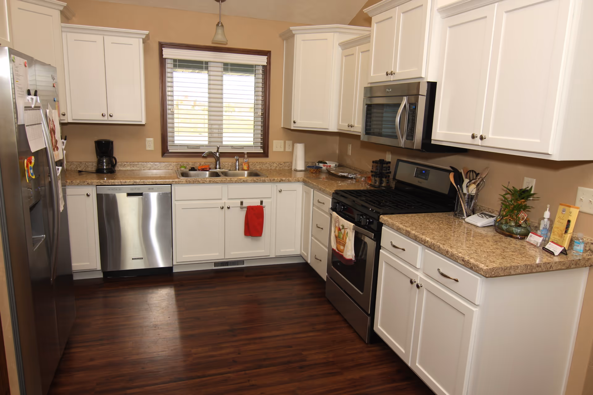 A modern kitchen with white cabinets, granite countertops, and stainless steel appliances including a refrigerator, dishwasher, stove, and microwave. There is a double sink under a window with blinds, a coffee maker on the counter, and various kitchen utensils and items on the countertops. The floor is dark wood.