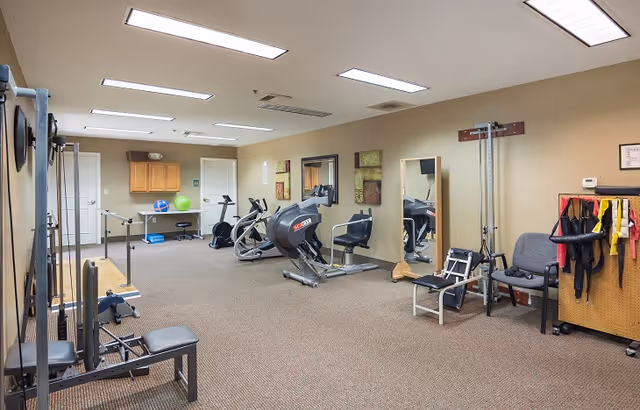 A fitness room in a senior living facility with various exercise equipment including stationary bikes, a weight machine, and resistance bands. The room has beige walls, carpeted floor, fluorescent ceiling lights, mirrors on the wall, and a small cabinet with exercise balls on top.