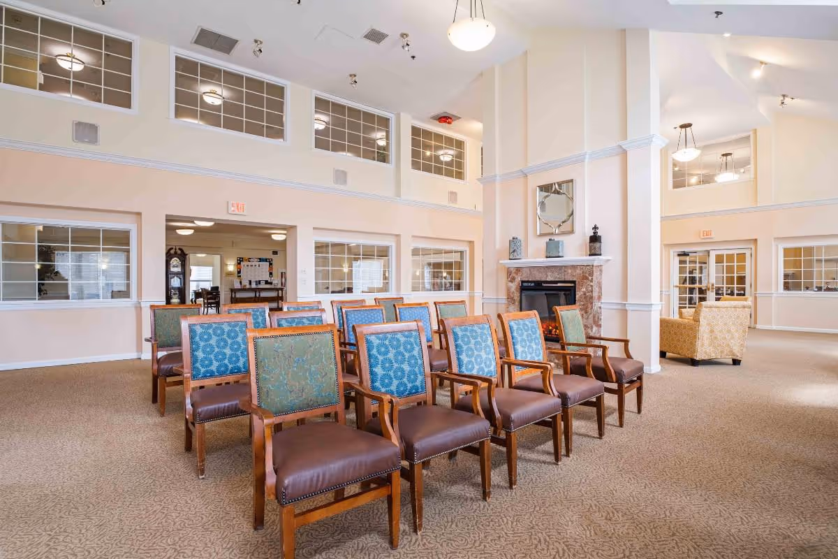 A spacious room with rows of wooden chairs featuring blue and green patterned upholstery, arranged facing a fireplace with a decorative mirror above it. The room has high ceilings with multiple windows near the ceiling, beige walls, and carpeted floors. There are additional seating areas with armchairs and visible doorways leading to other rooms.