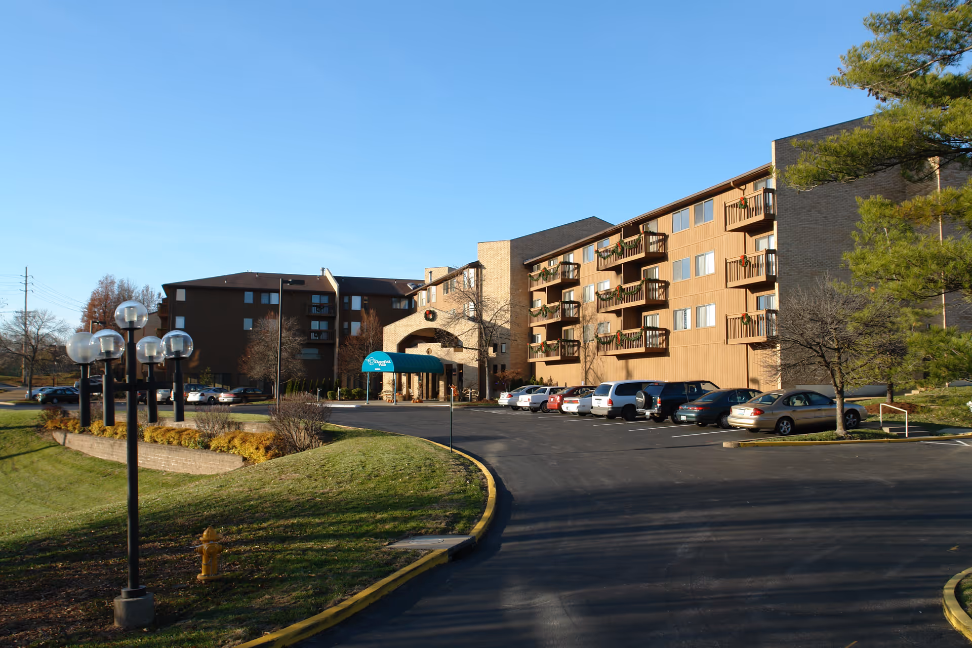 Exterior view of Chesterfield Villas senior living facility showing a multi-story building with balconies decorated with wreaths, a driveway leading to the entrance with a green awning, parked cars, and lamp posts along the grass area under a clear blue sky.