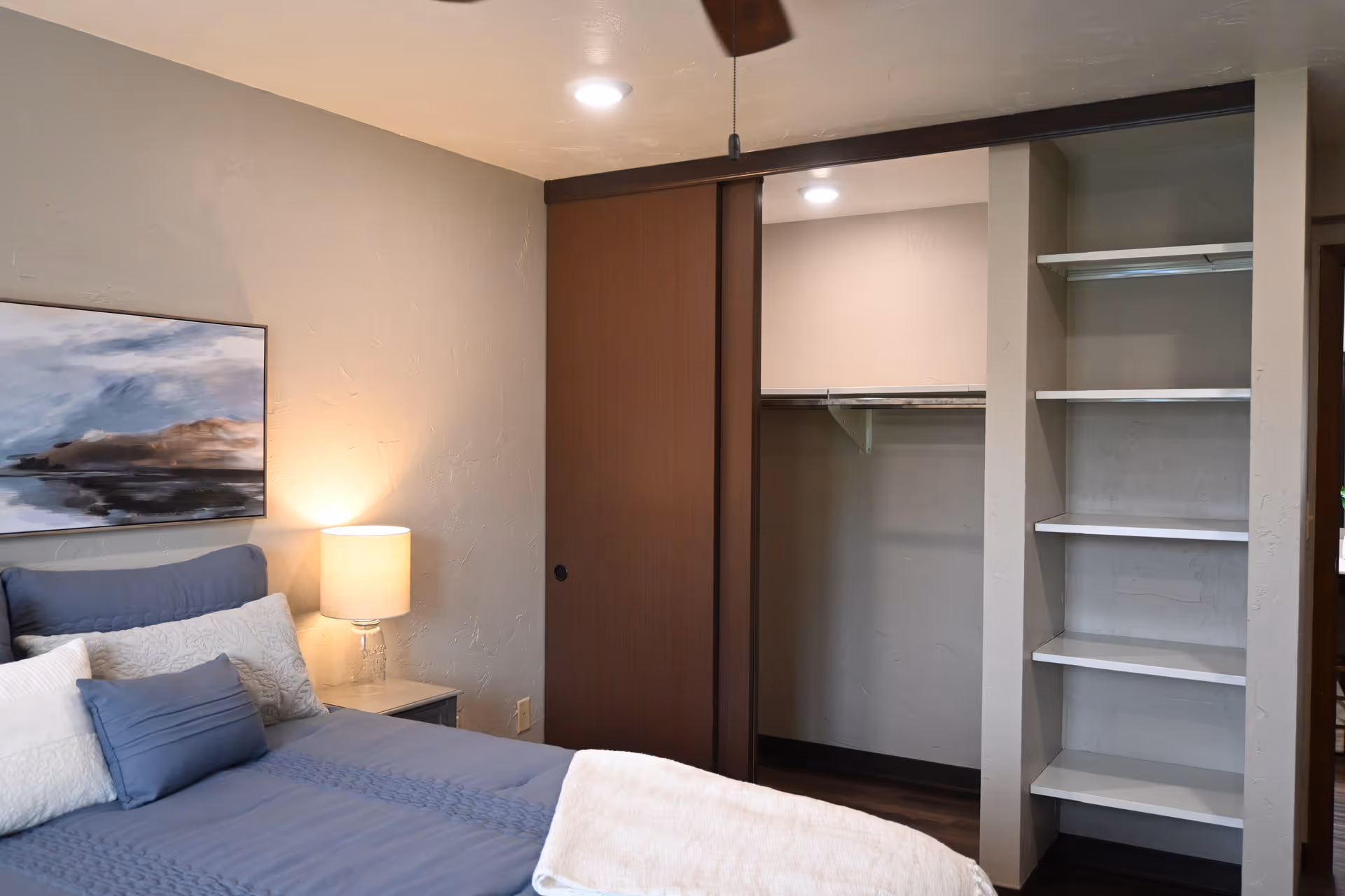 Bedroom with a made bed, bedside lamp and artwork, and an open closet with shelving and sliding door.