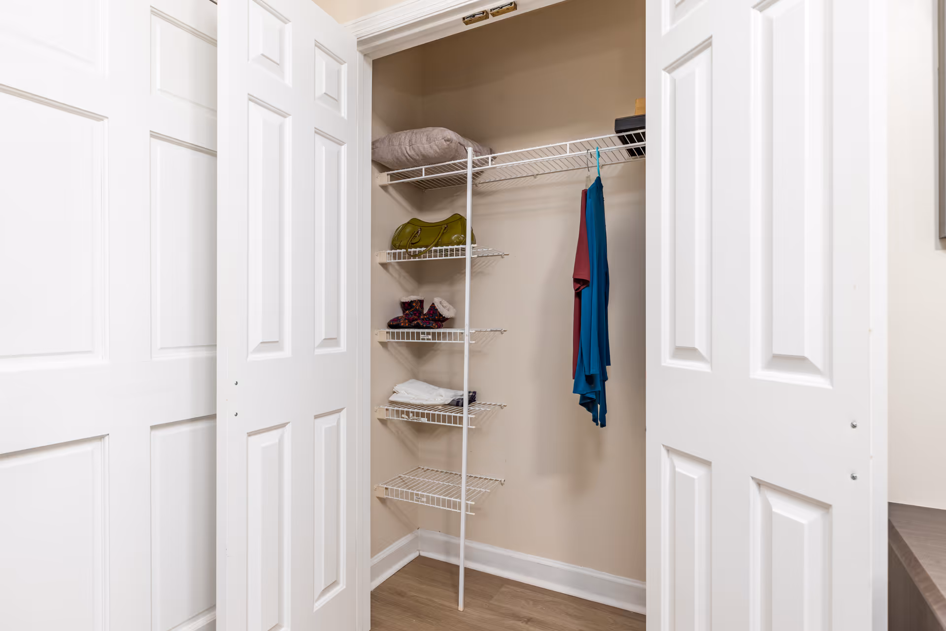 Open closet with white bi-fold doors showing wire shelving on the left side holding folded clothes, a green bag, and other small items, with two hanging garments on the right side against beige walls and wood flooring.
