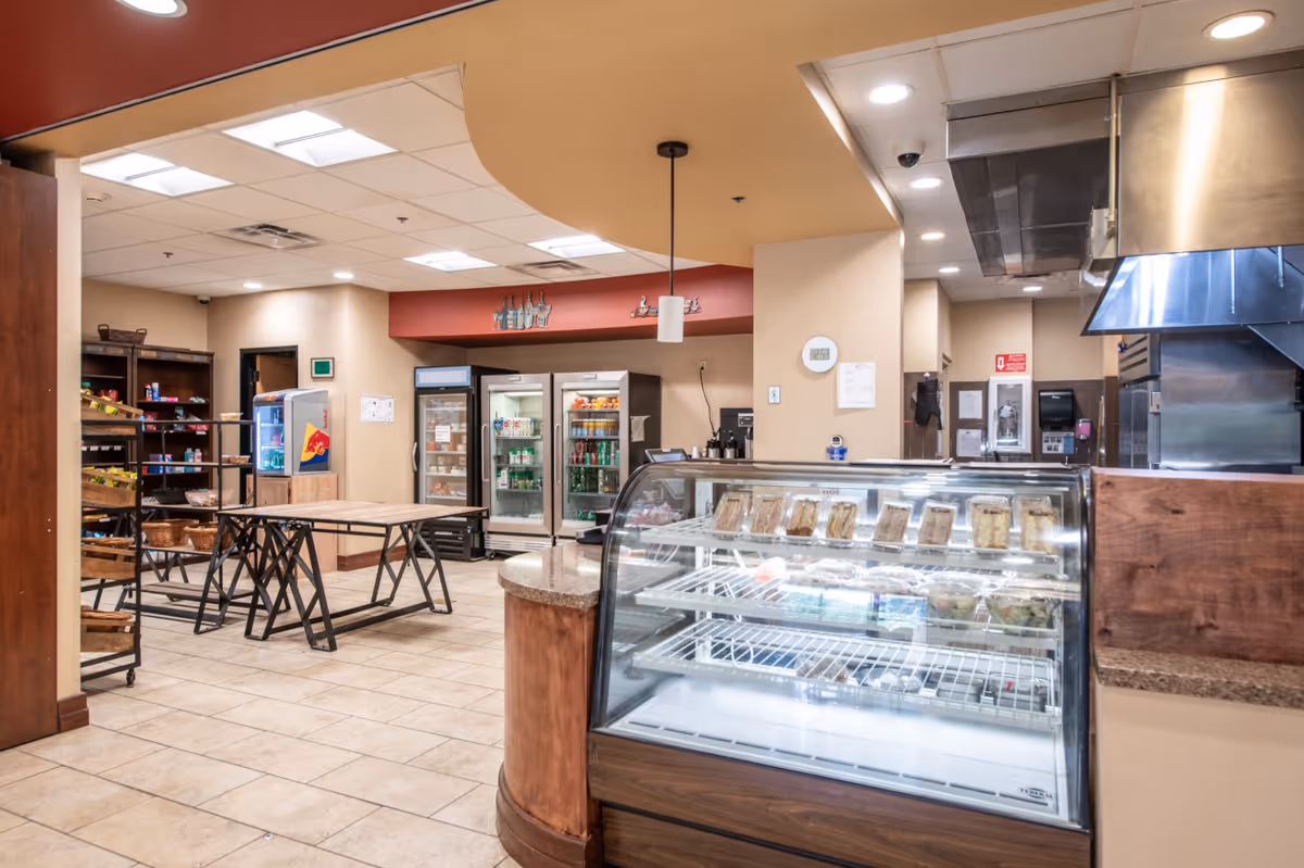 Interior view of a food service area in a retirement community featuring a glass display case with packaged food items, refrigerated beverage coolers, wooden shelving with baskets, and tables with metal legs under a beige ceiling with recessed lighting.