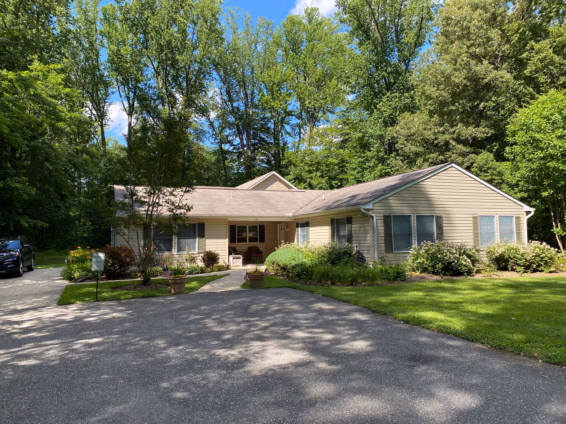 Single-story beige ranch-style building set among trees with a paved driveway, lawn, and landscaped shrubs.