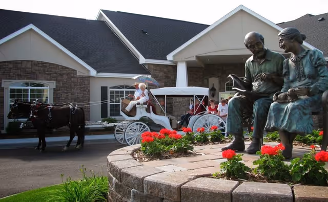 Bronze statues of an elderly couple and red flowers in a planter by the entrance of a senior living building, with a white horse-drawn carriage and people in the background.