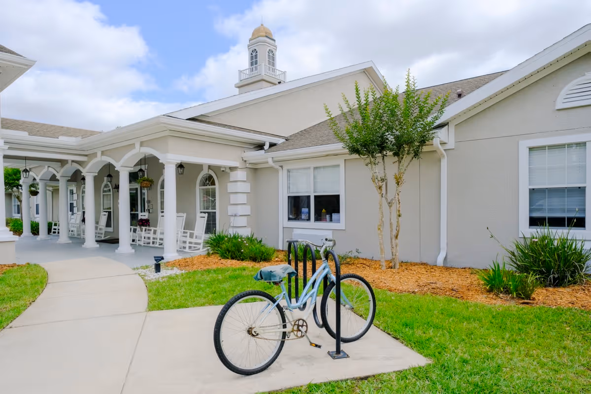 Front entrance of a light-colored senior living building with a covered porch, rocking chairs, and a bicycle parked at a rack on the walkway.