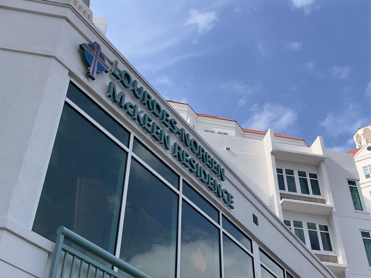Front exterior of the Lourdes McKeen Residence building with large windows and signage against a blue sky.