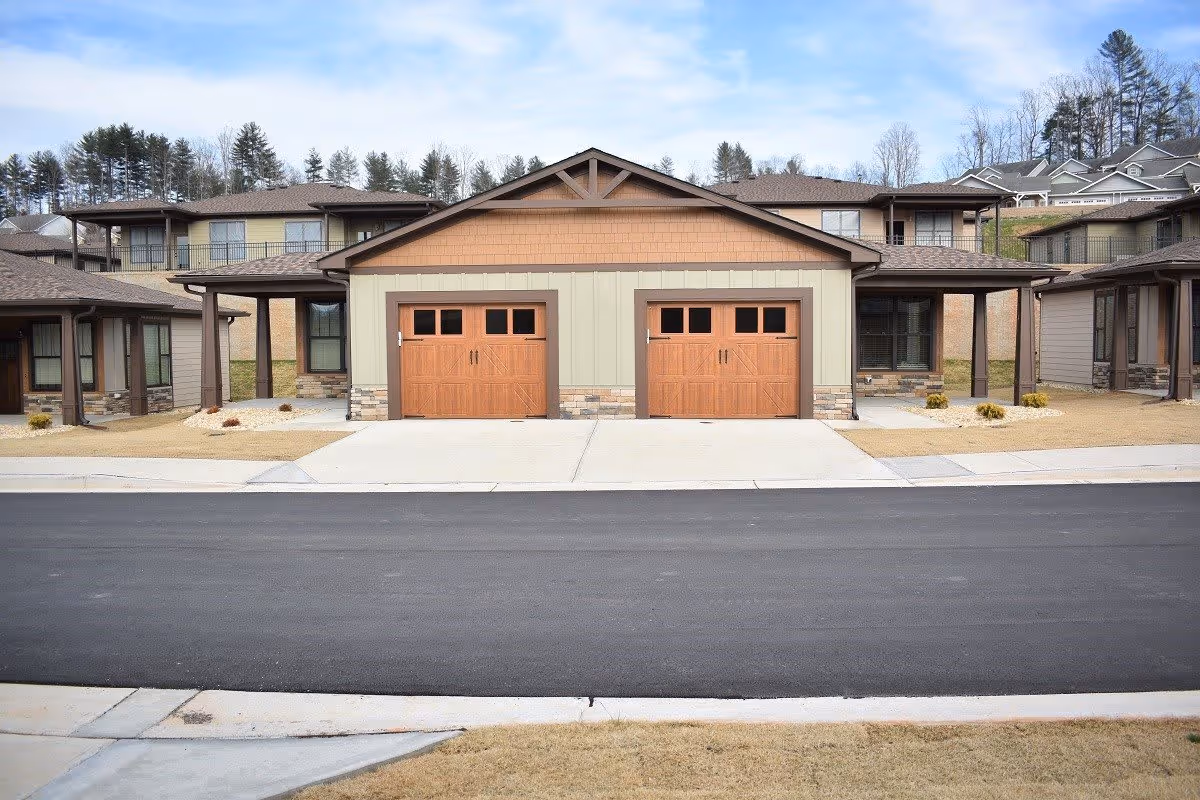 Exterior view of a residential building with two wooden garage doors in the center, surrounded by beige and brown siding. The building has a paved driveway and a black asphalt road in front. There are trees and other houses visible in the background under a partly cloudy sky.
