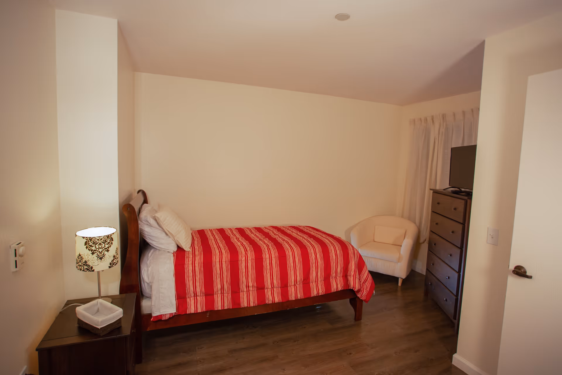 A bedroom with a single bed covered in a red and white striped comforter, a wooden nightstand with a decorative lamp, a white cushioned armchair, a dark wooden dresser with a TV on top, and cream-colored walls and curtains.