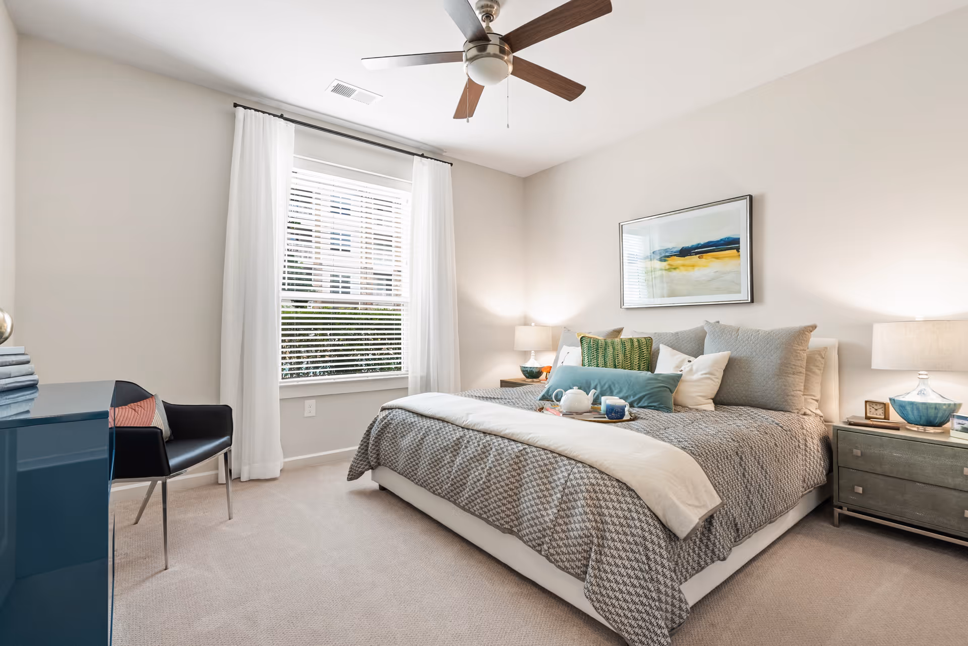 A bright and cozy bedroom with a large bed covered in a gray patterned comforter and multiple pillows. There is a tray with a teapot and cups on the bed. The room has beige walls, a window with white curtains, a ceiling fan, a blue dresser, a black chair, two bedside tables with lamps, and a framed abstract painting above the bed.
