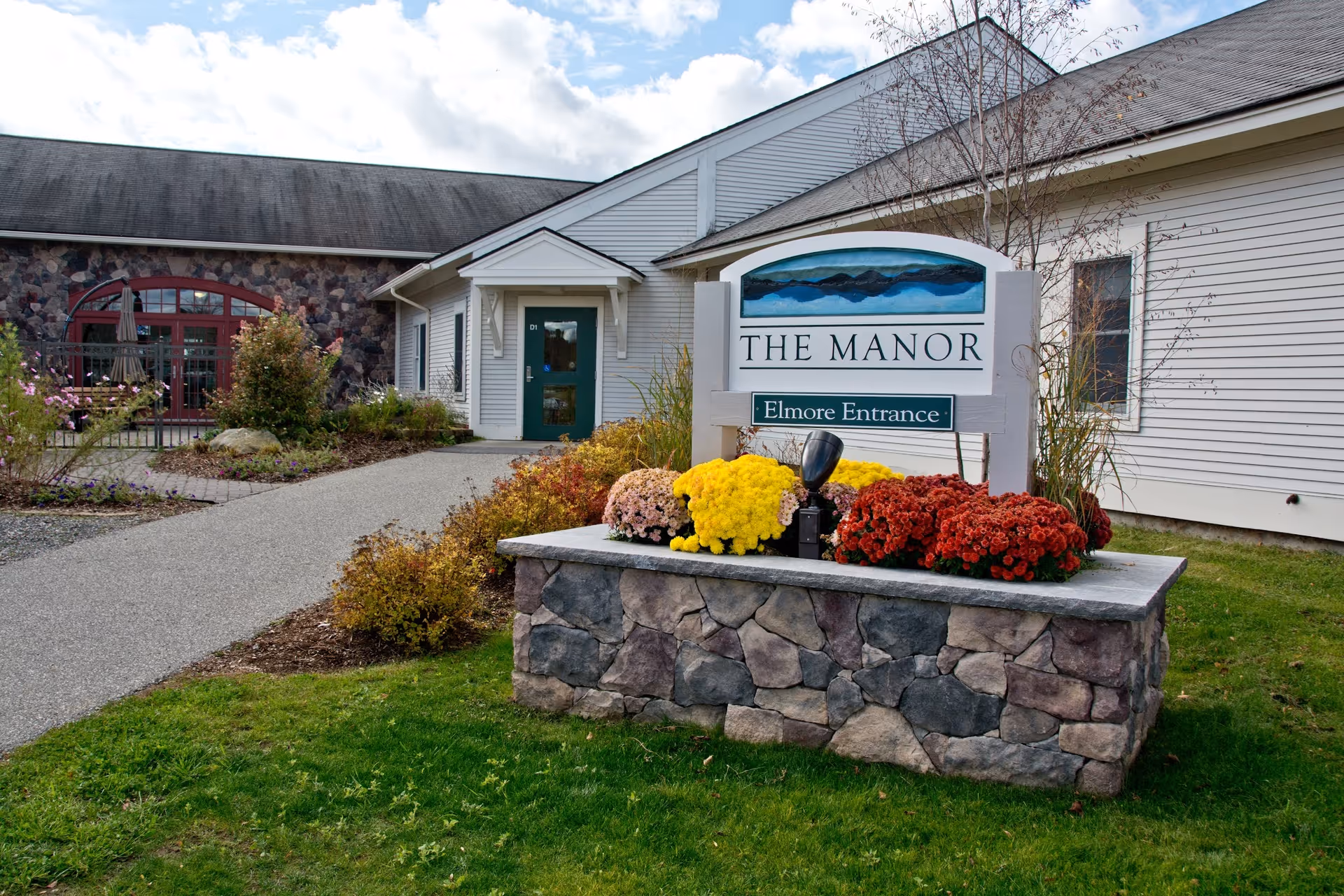 Entrance sign reading 'THE MANOR' and 'Elmore Entrance' in a flower-filled stone planter outside a single-story building.