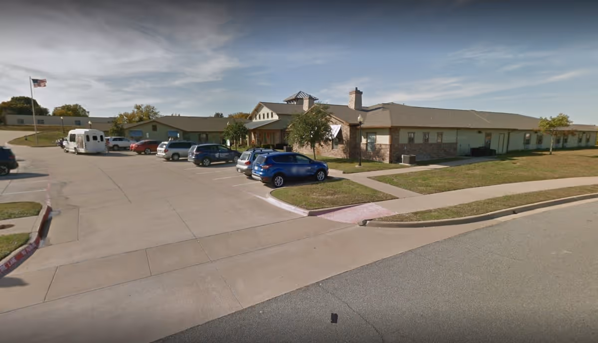 Exterior view of a single-story assisted living facility building with a parking lot in front. Several cars are parked, and an American flag is visible on a flagpole to the left. The building has a beige and brick facade with a sloped roof and a small covered entrance.