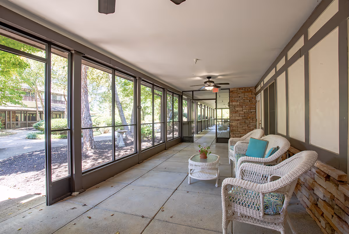 Screened covered walkway with wicker chairs, a small table with a potted plant, ceiling fans, and large windows overlooking trees.